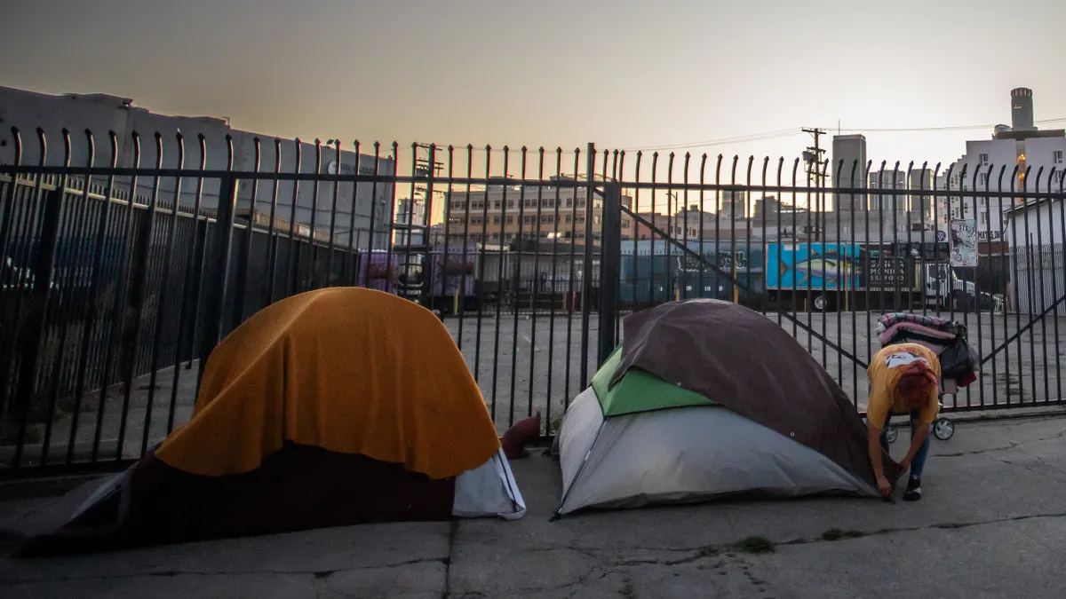 A person secures one of two tents set up in front of a tall gate with empty lot and buildings in the background.