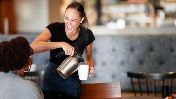 A person wearing black pours coffee into a white mug inside a restaurant. There is another person sitting at the table with her back to the camera.