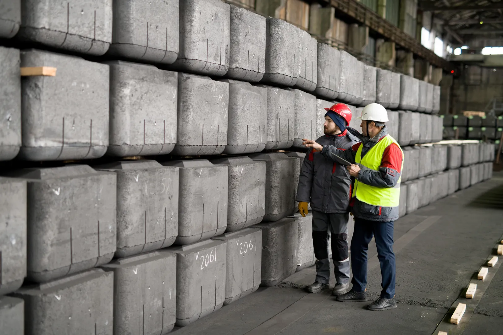 Two workers in safety gear conducting quality inspections of stacked large concrete squares inside an industrial plant.  