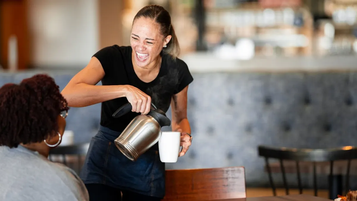 A person wearing black pours coffee into a white mug inside a restaurant. There is another person sitting at the table with her back to the camera.