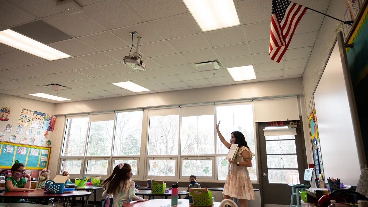 An adult stands at the front of a classroom with arm in the air. Students sit at desks in the classroom looking at adult.