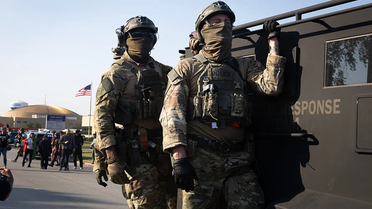 Federal law enforcement agents in uniform with masks covering their faces ride an armored vehicle through demonstrators during a protest outside an immigrant processing and detention center.