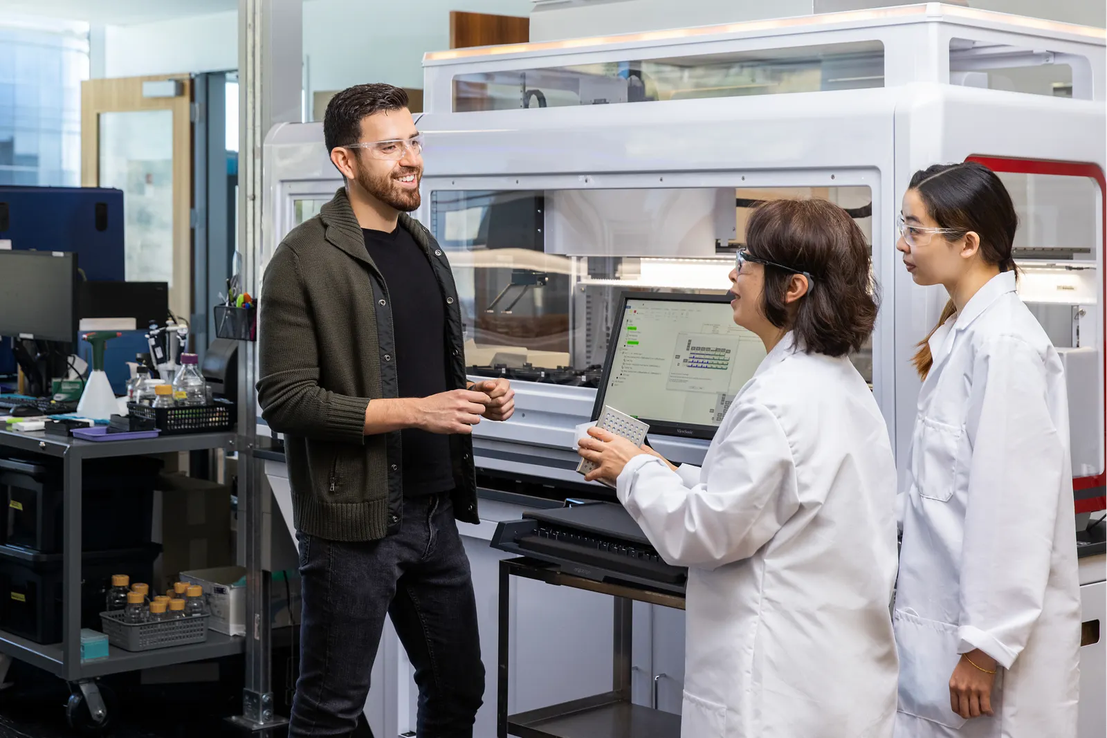 Arturo Elizondo talks to a pair of people in lab coats and safety goggles in a lab. Large pieces of equipment, computers and small bottles are in the background.