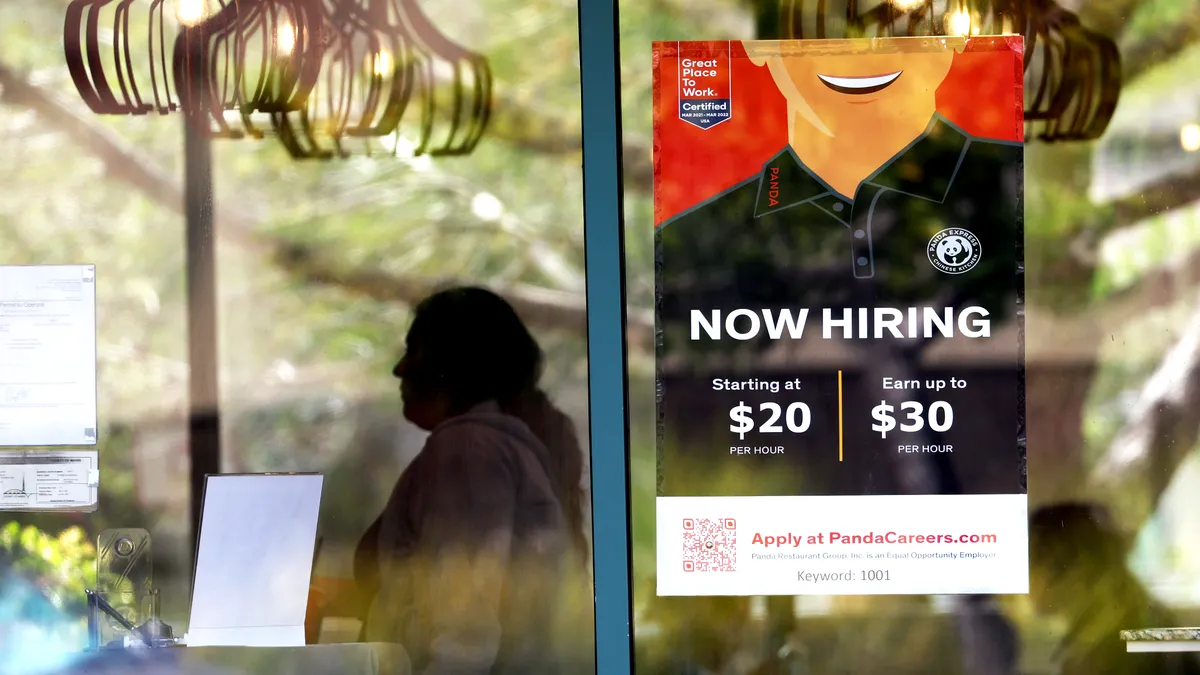 A "now hiring" sign is posted in a restaurant window, advertising an hourly pay rate of $20 to $30 per hour.