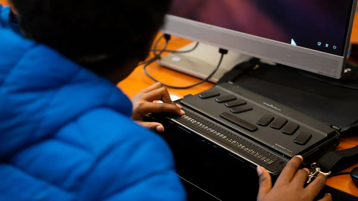 A student, whose  part of hand and part of head can bee seen, is using a reading device at school. The device is black and has buttons.