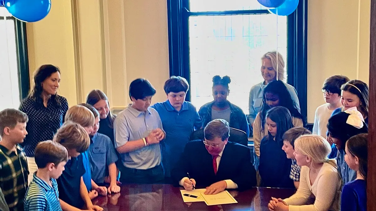 A group of elementary students and adults stand around an adult seated at a desk. That seated person is writing on a paper. The group is in a room with windows and blu balloons.