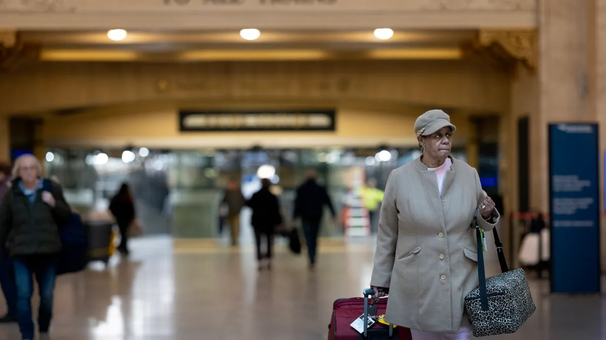 A woman in a gray hat and jacket walks toward the camera against a blurred background of people and a sign reading "To All Trains."