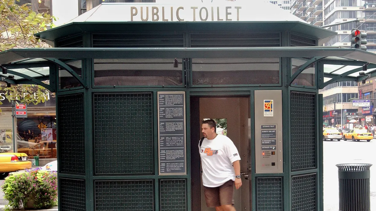 A person wearing a tee shirt, sneakers and shorts walks out of a public toilet.