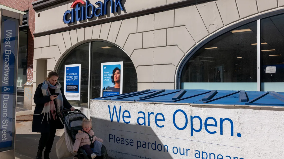 A person with a kid in a stroller walks past a bank branch while looking at their phone.