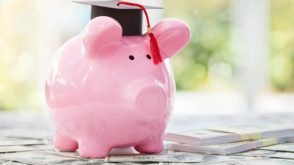 A pink piggy bank with a black graduation cap on top of its head stand on top of stacks of bills against an outdoor sunny background.