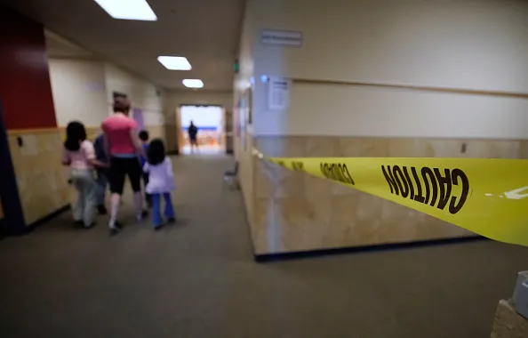 An adult and small children are walking away from the camera down a hallway in a school building. In the forefront is yellow caution tape across another hallway.