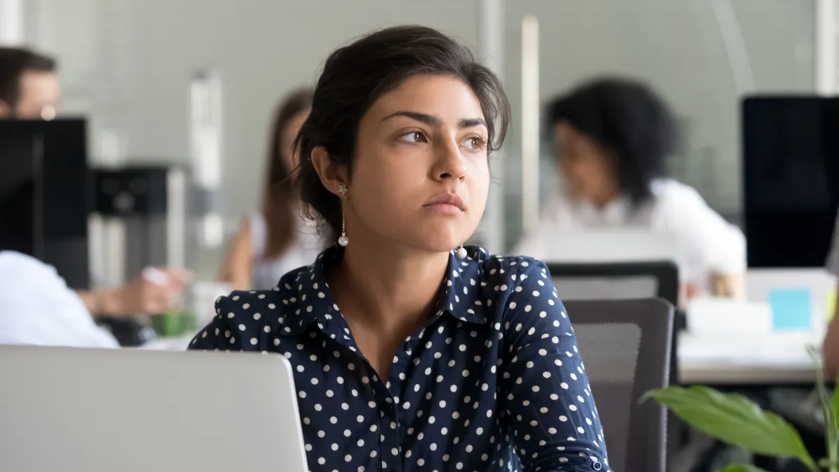 An office worker in a polka-dotted shirt sits at her computer and looks thoughtfully in the distance.