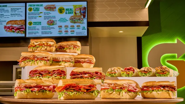 A photograph of 15 various sandwiches on a table with a Fresh Value Menu highlighted in the background from Subway
