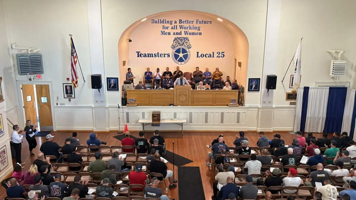 A view looking down at a union hall from a balcony. Behind the stage on the wall are the words "Teamsters Local 25" and the IBT logo. People are gathered on stage and seated in the audience.
