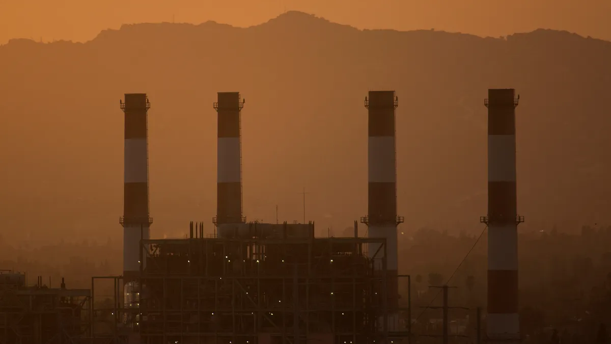 A gas-powered power generation plant is seen with four red and white striped pillars rising from the facility with a mountain's outline visible through a orange haze.