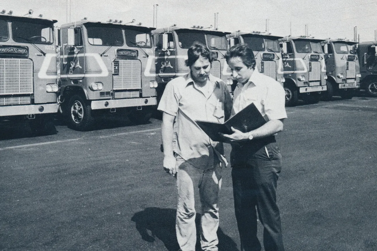 Two people review a binder in front of a row of seven Saia Freightliner trucks in an undated, black-and-white photo.