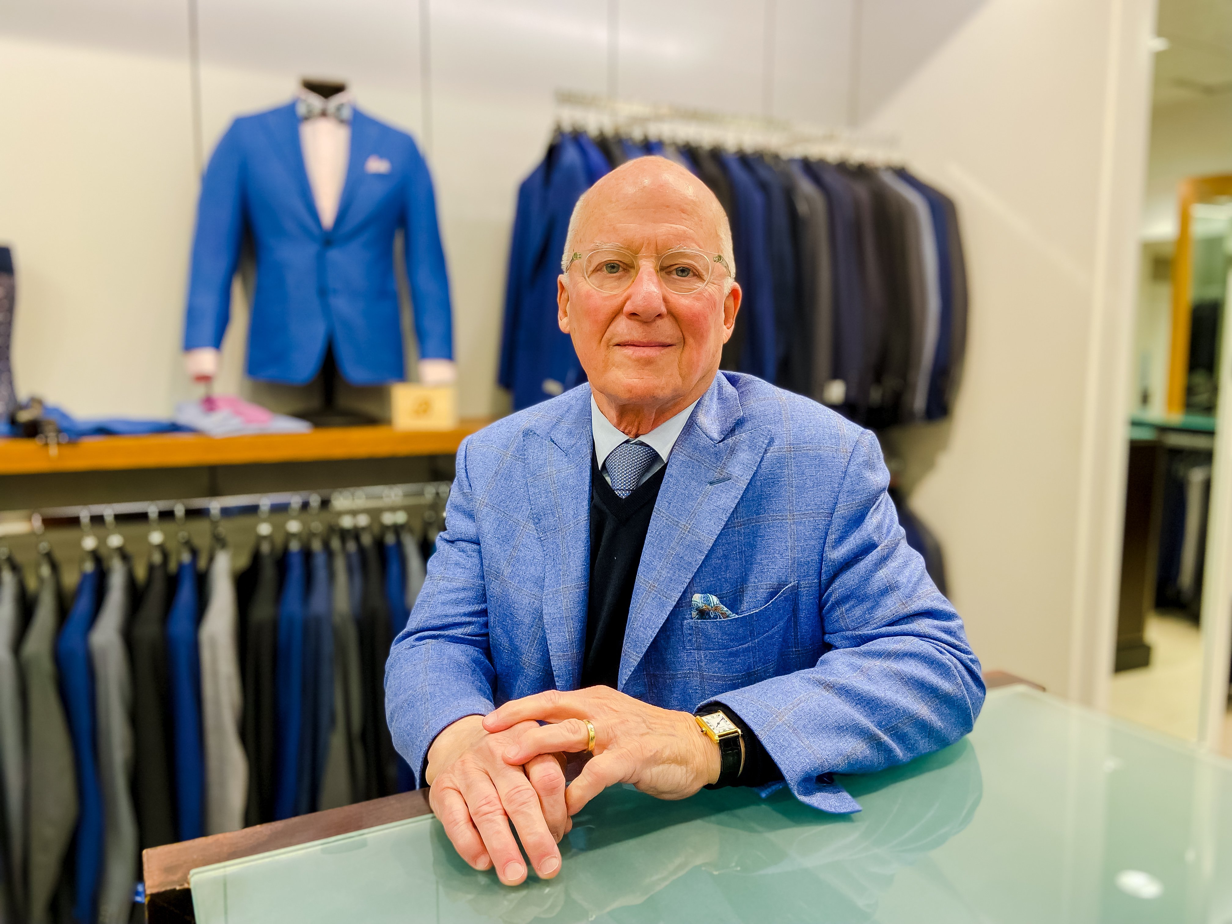 A person in a blue suit sits with hands folded, with displays of clothing in the background.