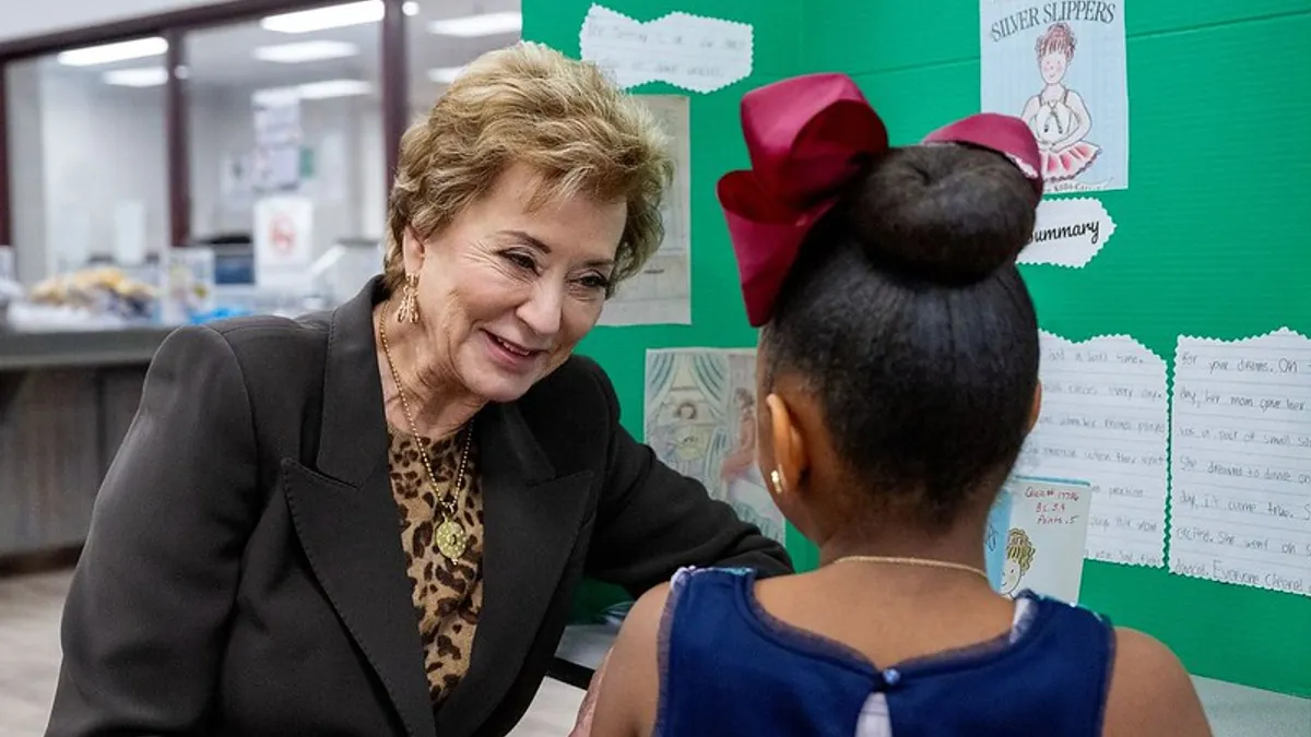 An adult sits at a table near a wall in a classroom. A student is sitting across from McMahon with their back to the camera