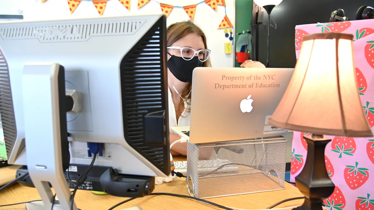 A person sits at a desk behind two computer monitors. The person is looking at a monitor and wears eyeglasses