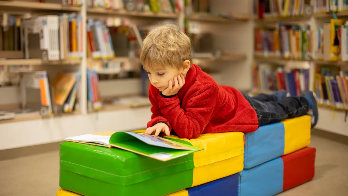 A young students lies on stomach on top of colorful cushions. The student, who is in a room with bookshelves,  is looking down at a picture book.