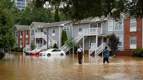 Two people walk through knee-deep brown flood water in front of a two-story apartment complex, with a taller apartment building in the background.