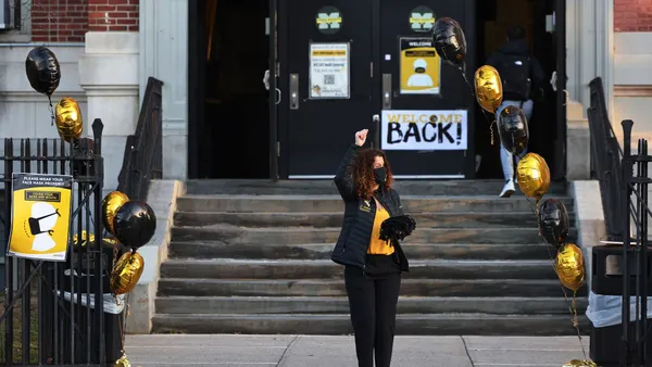 An adult stands outside a school building with their arm in the air. There are balloons and signs near the school entrance.
