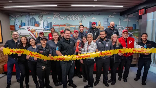 A group of people stand behind a yellow ribbon while a person in the middle holds up large scissors that are about to cut the ribbon.