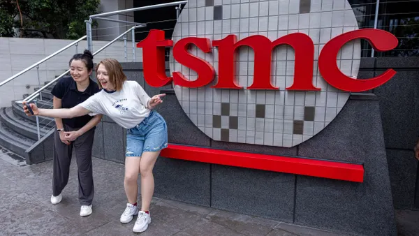 Two people taking a selfie with a cell phone in front of the red TSMC logo.