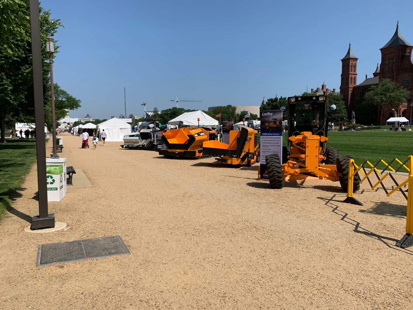 On a bright, sunny day, a line of construction equipment is present from the foreground to the background. They are positioned on gravel, with green grass behind them.