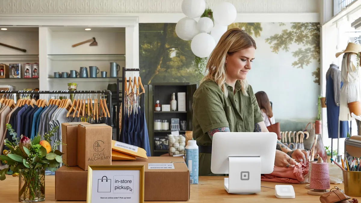 A person at a table in a clothing store wrapping up a piece of garment with a mobile payment stand with the Square logo in from of them.