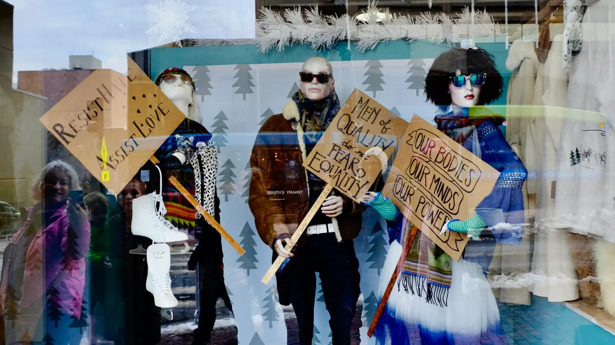 A resale boutique window features mannequins wearing secondhand clothing and holding handwritten signs touting civil rights.