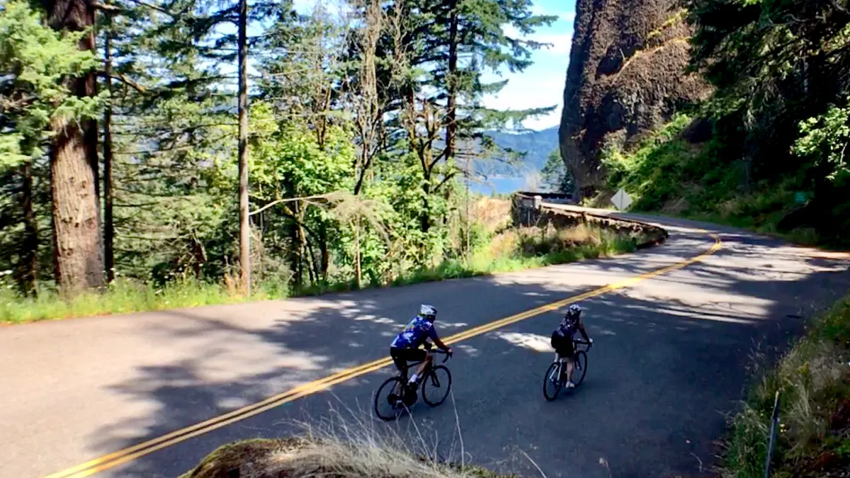 Two people on bicycles along a two-lane road surrounded by trees.