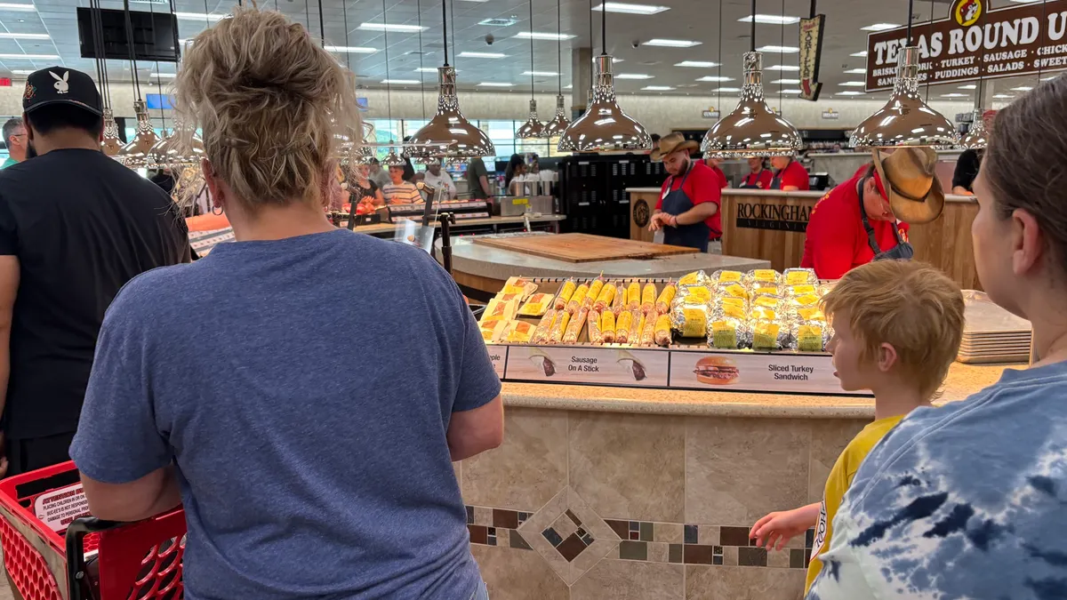 A photo of the interior of a large store. People are waiting in line to get sandwiches which are being kept warm under heat lamps.