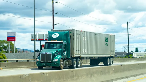 An R+L Carriers semi-truck on a highway hauling an intermodal cargo container with a concrete barrier in the foreground.