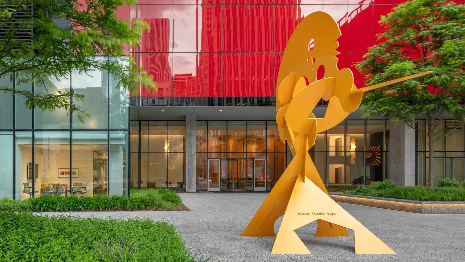 A yellow metal abstract sculpture in front of a red building.