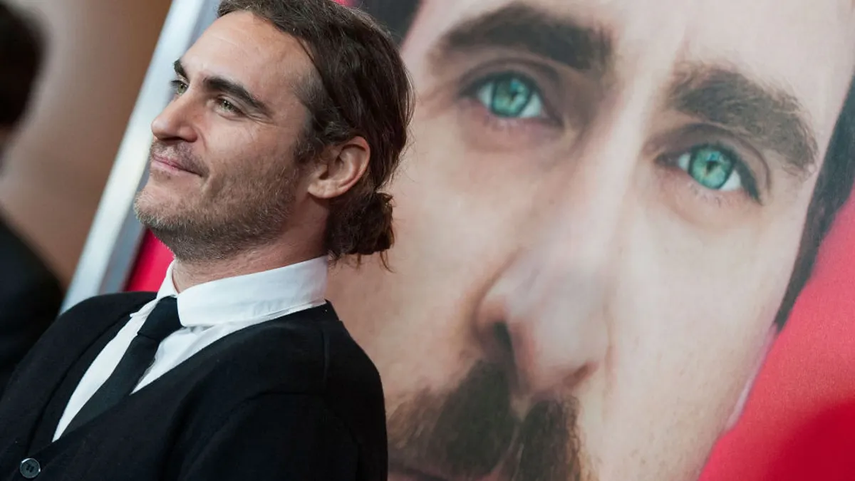 Actor Joaquin Phoenix stands smiling in front of his own face on the poster for the film "Her."