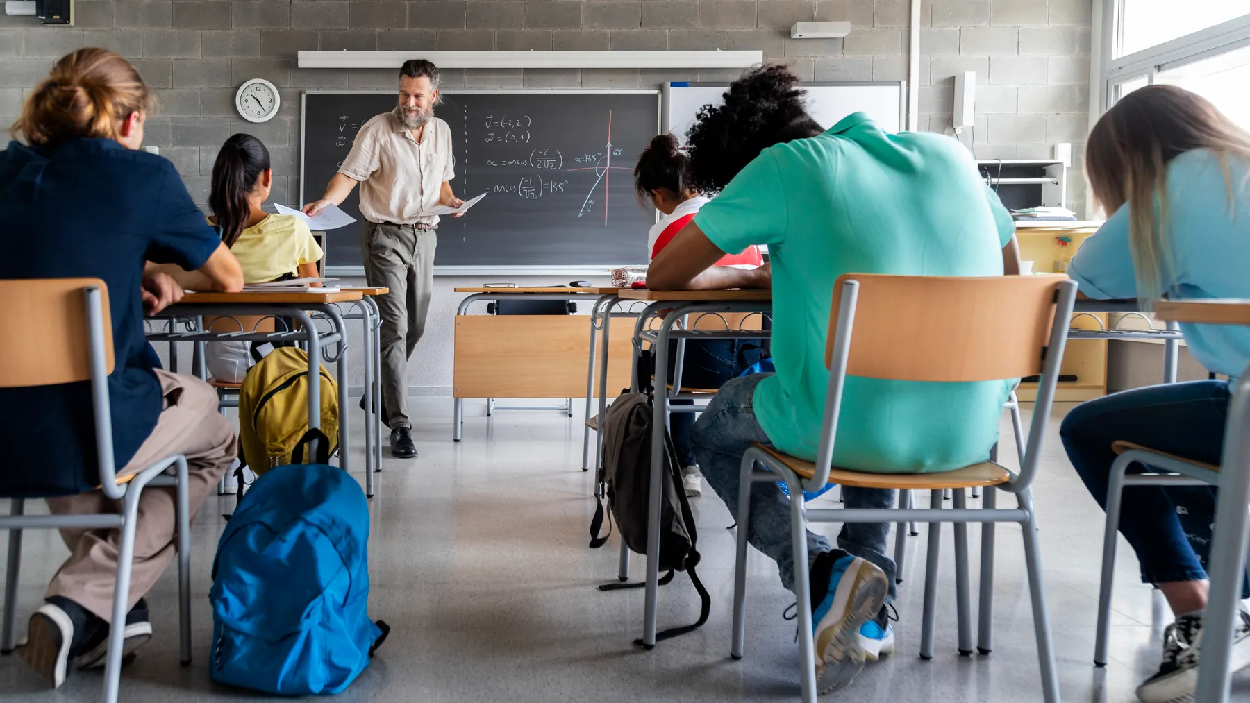 Students sit in desks in a classroom facing a backboard and a teacher who is handing out papers.