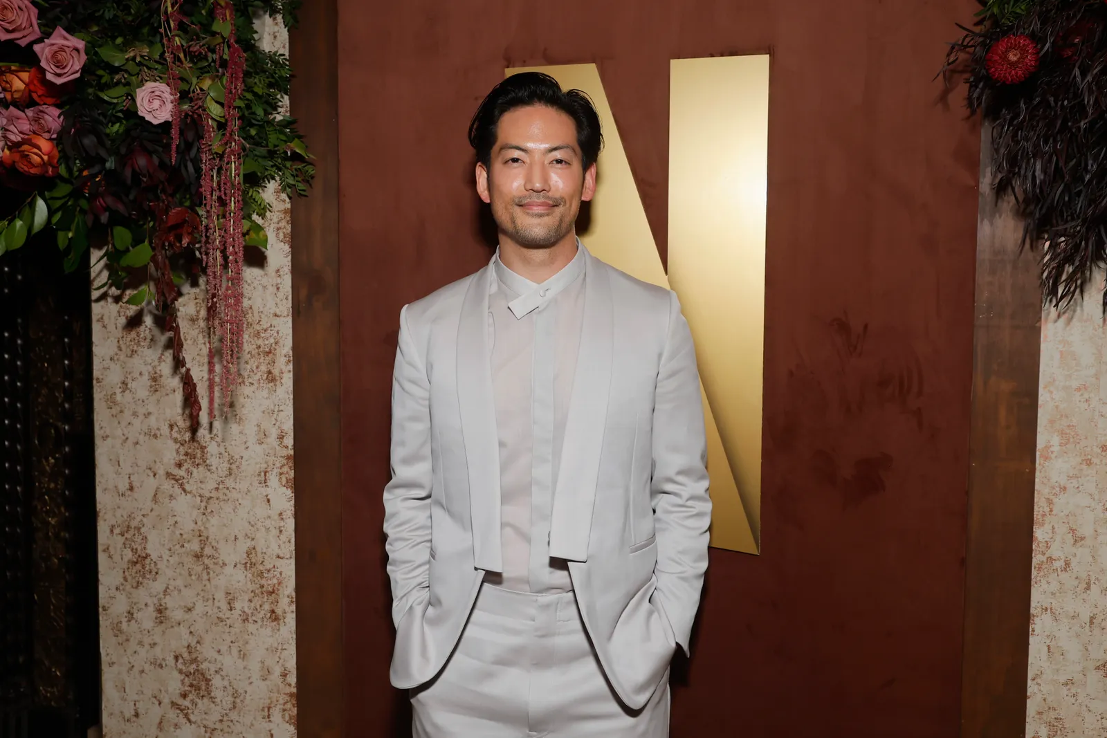 A person in a grey suit with a sheer shirt poses for the camera and smiles.