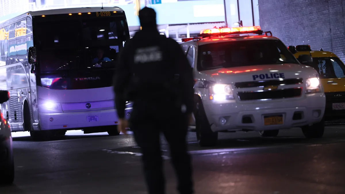 A bus is behind a police car in a large garage. At the center of the photo is a uniformed police officer with back toward camera.