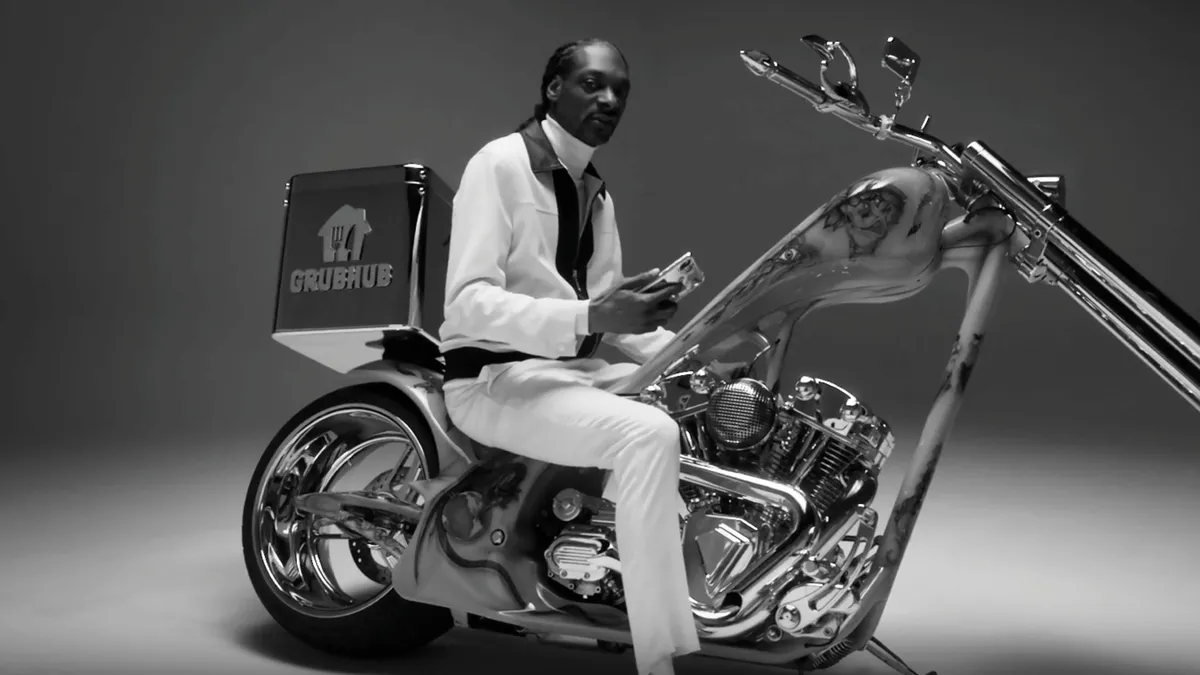 A black and white photo of Snoop Dogg sits atop a chrome motorcycle with a Grubhub container on the back.