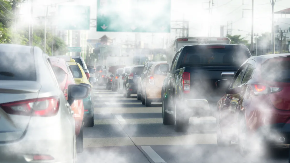 Bumper-to-bumper cars driving away from the camera spewing gray exhaust gases.