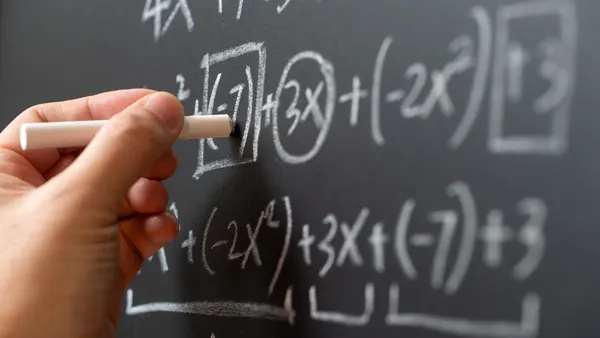 A hand is holding a piece of chalk. The chalk is writing algebraic formulas on a chalkboard
