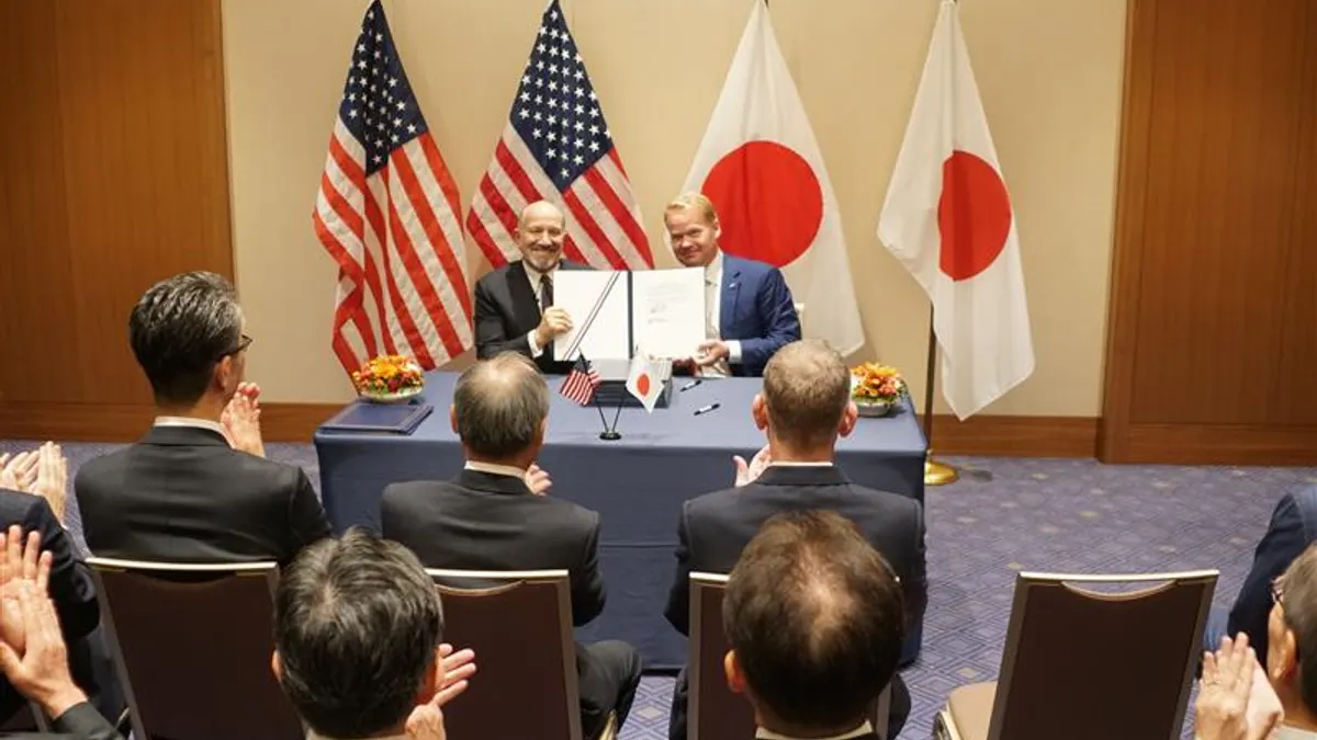 Two men at a table hold up a signed document before American and Japanese flags.
