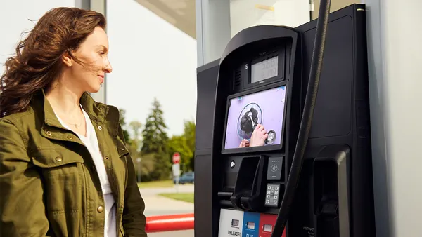 A woman stands at a gas station, looking at a modern fuel pump with a digital screen while holding the fuel nozzle, with trees and a roadway visible in the background.