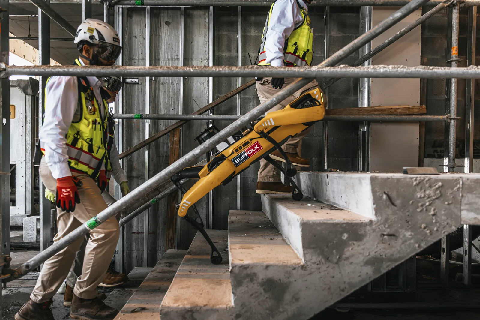 two people in personal protection gear make their way up a set of stairs while a four-legged robot walks beside them.