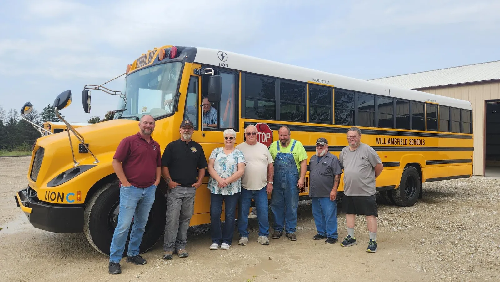 A group of adults stand in front of an electric school bus. One person is sitting inside the bus in the driver&amp;#x27;s seat