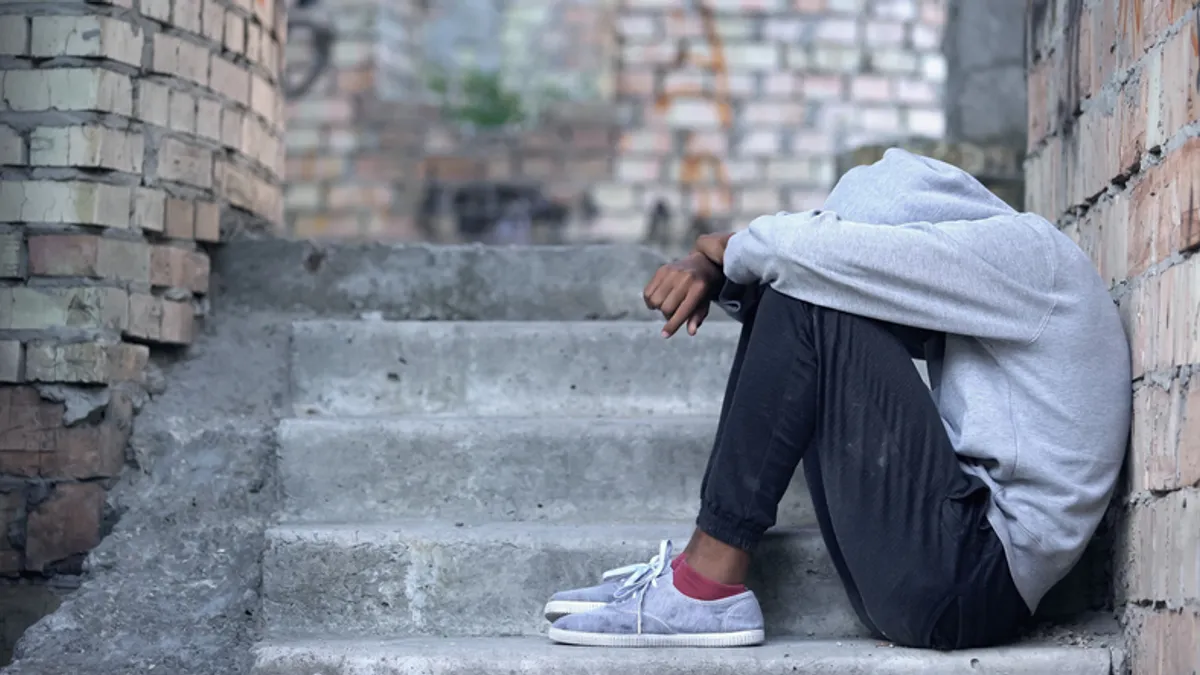 A person is sitting outside on steps. The person's body is leaning against a wall. Their sweatshirt covers their head and their head is bent down toward their knees.