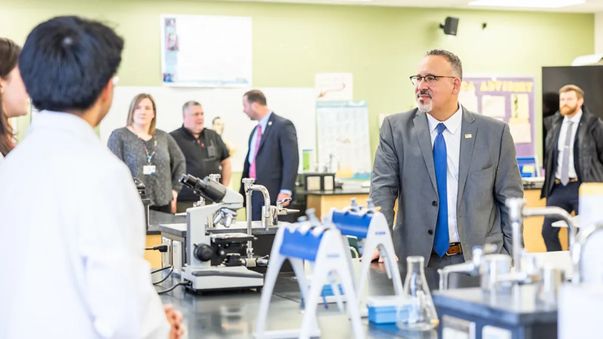 A person is standing at a side angle in a classroom with laboratory tables facing another person whose face is turned to the other person. Other people are standing around in the background