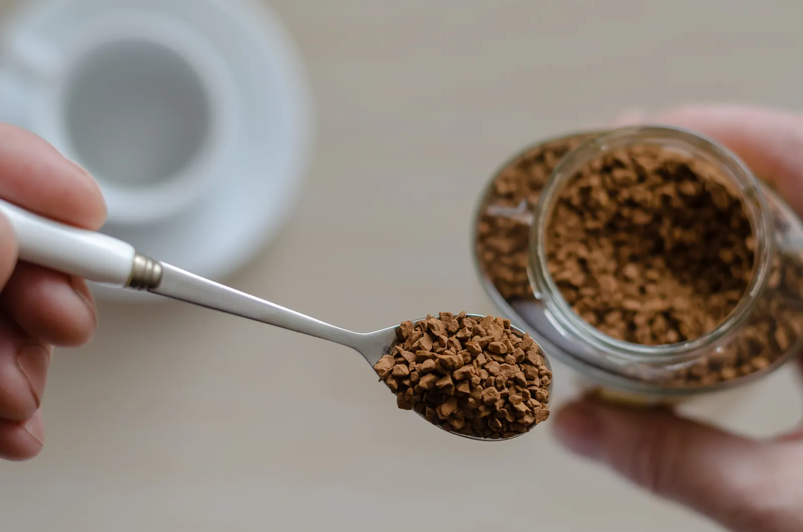 Overhead view of a person holding a spoon of instant coffee in one hand and the container of it in the other. A white coffee cup is blurred and sitting on a surface.