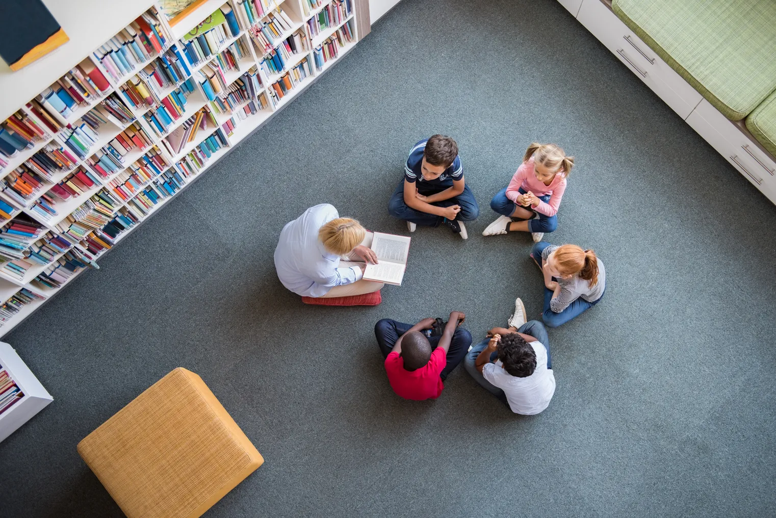 An above camera shot of an adult and a few students sitting on the floor in a circle in a library. The adult has a book open.